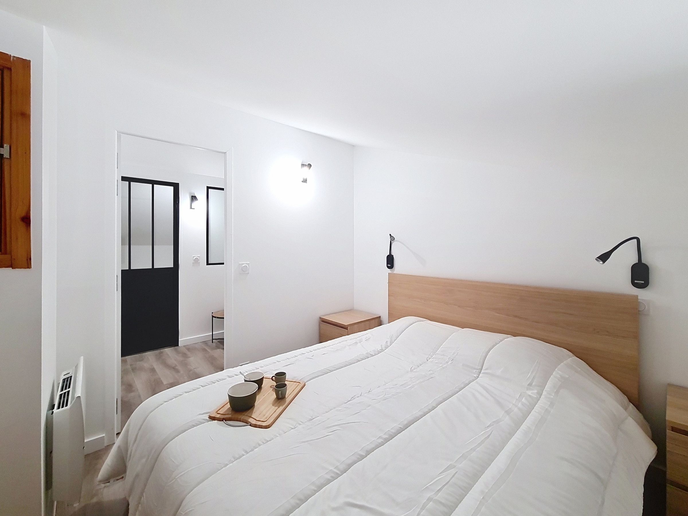 Een slaapkamer met een bed, nachtkastje en een bijzettafeltje. Er staat een klein houten krukje en een dienblad met bekers en een schaal.