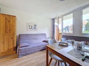 A cozy room with a wooden wardrobe, a cushioned sofa, a dining table with chairs, and a window with a view of buildings. - Cozy studio with balcony, ski-in ski-out (Les Menuires)