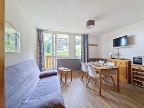 A room with a sofa, dining table, chairs, a TV, and a window with curtains. - Cozy studio with balcony, ski-in ski-out (Les Menuires)