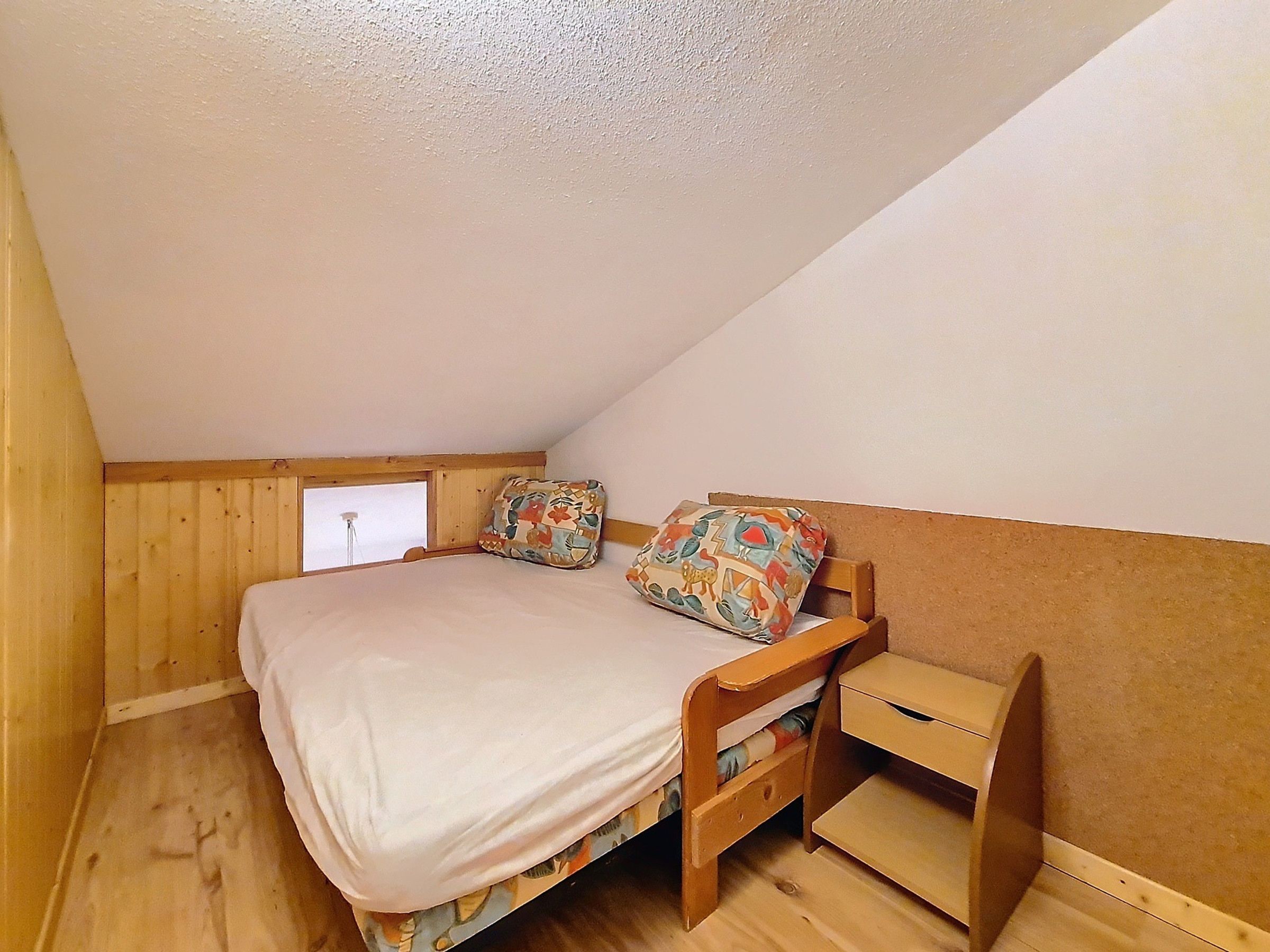 Ein kleines Zimmer mit einem Einzelbett, geneigter Decke, Holzwänden und einem Fenster.