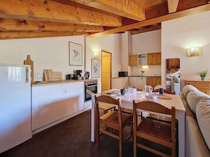 A compact kitchen with white appliances, wooden ceiling beams, a dining table with chairs, and a sofa in a cozy living area. - 3-room apartment, southwest balcony, at the foot of slopes (Les Menuires)