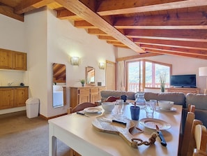 A dining area with a table set for two, wooden beams on the ceiling, a television in the background, and a view of the outdoors through large windows. - 3-room apartment, southwest balcony, at the foot of slopes (Les Menuires)