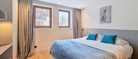 A bedroom with a bed, two windows, a lamp, and a painting on the wall.