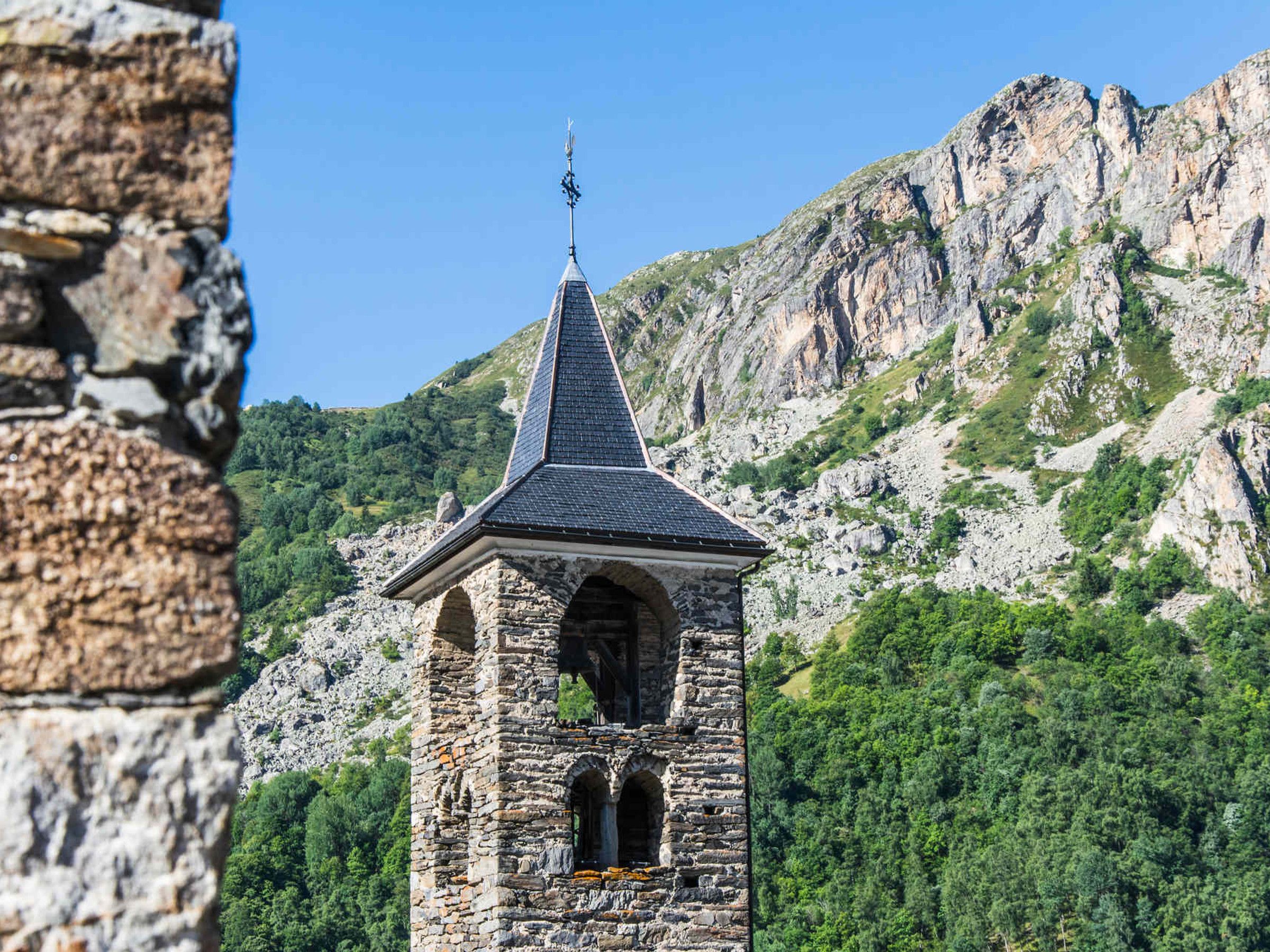 Une tour de pierre, surmontée d’un toit en forme de flèche et d’une girouette, se dressant sur un fond de montagnes.