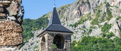 Une tour de pierre, surmontée d’un toit en forme de flèche et d’une girouette, se dressant sur un fond de montagnes.