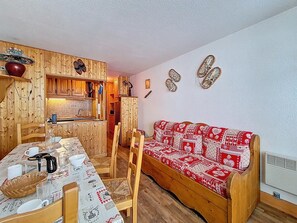 Une pièce chaleureuse avec une table à manger en bois, des chaises, un canapé à motifs rouges et blancs, et un coin cuisine doté de meubles en bois et d’un porte-chaussures suspendu.