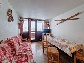 A cozy living room with a red patterned sofa, a dining table with a white tablecloth, and a television on a wooden stand. - Spacious apartment - 6 guests at the foot of the slopes (Les Menuires)