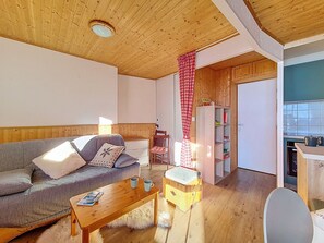 A cozy living room with a wooden ceiling, a sofa with cushions, a coffee table, a small wooden stool, and a kitchen area with a shelf and a cabinet. - Bright apartment for 5. Close to slopes. Balcony. (Les Menuires)