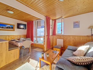 A cozy room with a wooden ceiling, a dining area with a table and chairs, a sofa with a cushion, and a television on the wall. - Bright apartment for 5. Close to slopes. Balcony. (Les Menuires)
