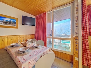 A dining area with a table set for two, a television mounted on the wall, and a view of a snowy mountain through a large window. - Bright apartment for 5. Close to slopes. Balcony. (Les Menuires)