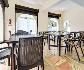 Un comedor amplio con varias mesas y sillas, ventanas grandes con contraventanas de madera, y un cartel de seguridad contra incendios en la pared.