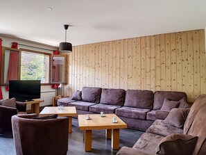 A modern living room with a wooden panel wall, a large sofa, a TV, and a coffee table. - Spacious 7-room apartment near slopes with sauna, hammam, and wifi (Les Menuires)