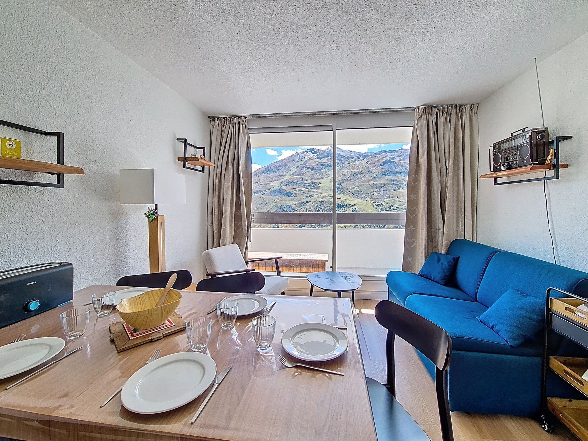 A room with a dining table set for four, a blue sofa, a television, and a view of mountains through large windows.