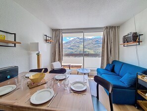 A room with a dining table set for four, a blue sofa, a television, and a view of mountains through large windows. - Renovated apartment 5 pers. Ski-in/ski-out, south balcony, WIFI (Les Menuires)