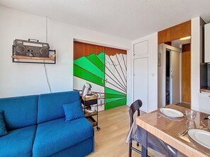 A compact living space with a sofa, a dining table with chairs, a kitchen area, and a wall with a colorful mural. - Renovated apartment 5 pers. Ski-in/ski-out, south balcony, WIFI (Les Menuires)