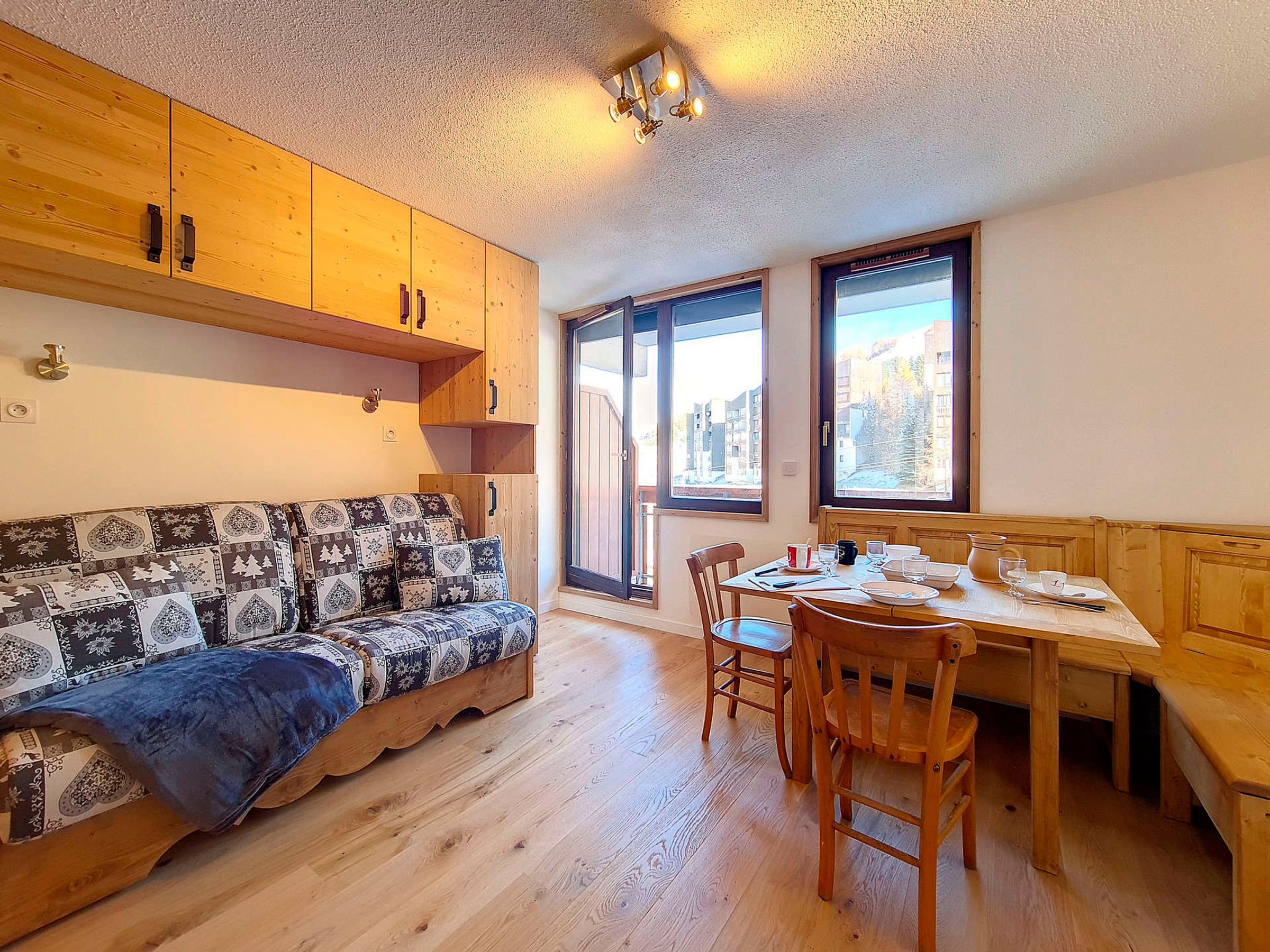 Een gezellige kamer met een houten bank, een eettafel met stoelen en een groot raam met uitzicht op gebouwen en bomen.