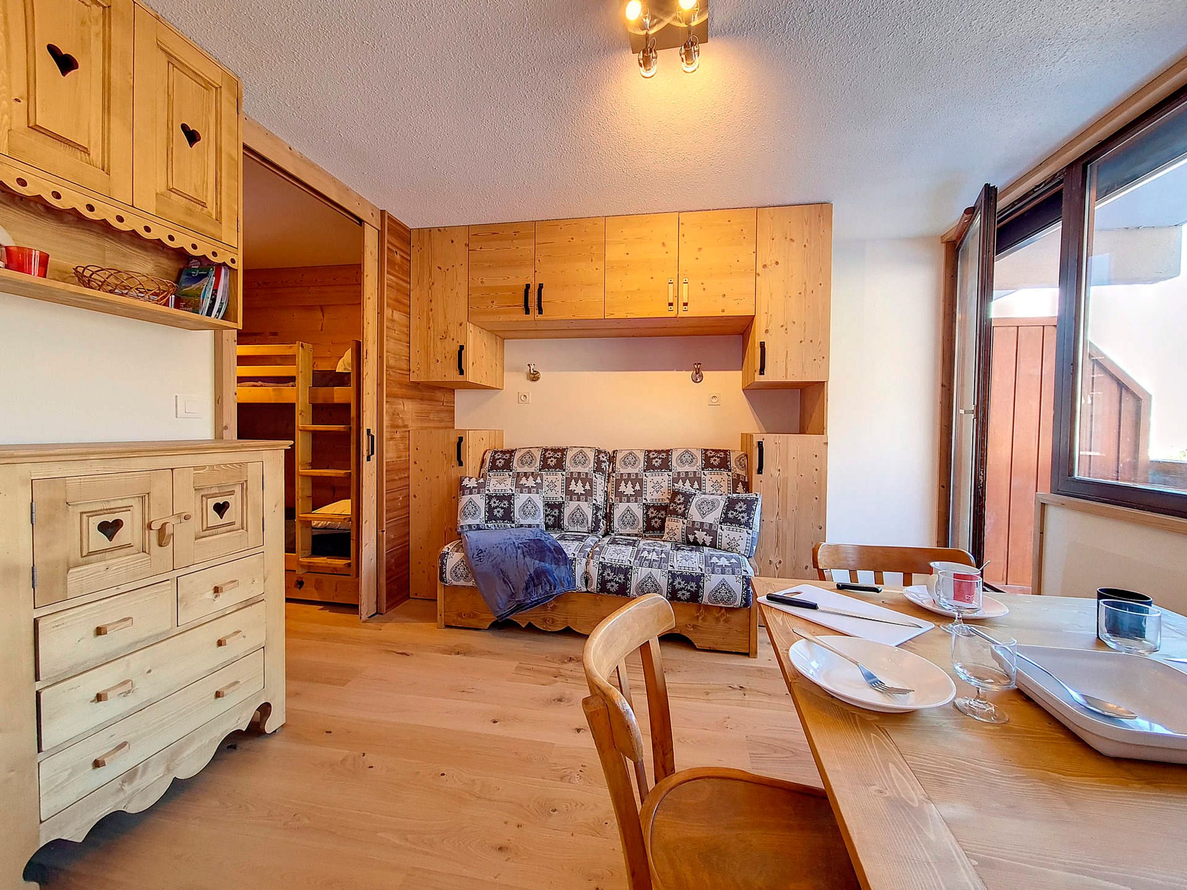 A cozy room with wooden furniture, a bed with patterned bedding, a dining table set for two, and a window providing natural light.