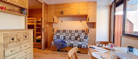 A cozy room with wooden furniture, a bed with patterned bedding, a dining table set for two, and a window providing natural light.