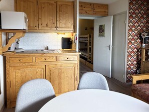 A compact kitchen with wooden cabinets, a microwave, and a sink. There is a dining area with a table and two chairs. - Cozy studio in the heart of the resort - 4 guests (Les Orres)