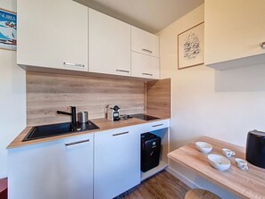 A compact kitchen with white cabinets, a wooden backsplash, a built-in dishwasher, and a small coffee table with cups. - Renovated studio at the slopes with WIFI (Les Menuires)