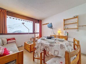 A cozy dining area with a table set for two, a wooden chair, a small wooden cabinet, a shelf with glassware, and a window with a view of a snowy landscape. - South-facing balcony apt - 4 guests (Les Menuires)