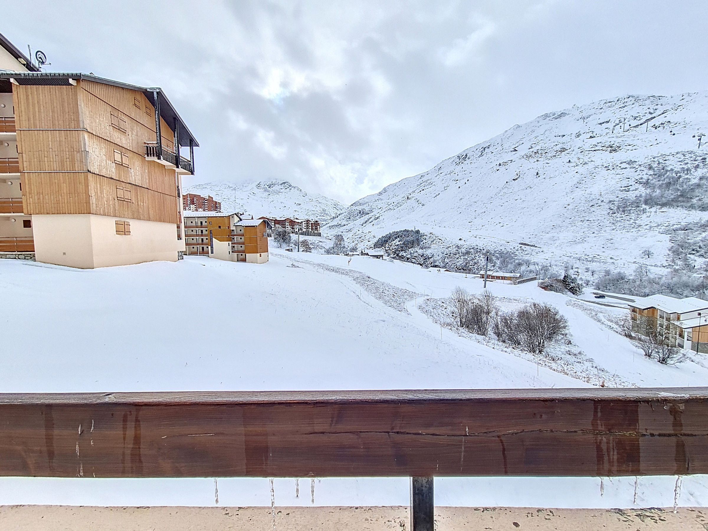 A snow-covered mountain landscape with buildings, including a multi-story structure with balconies, and a wooden fence in the foreground.