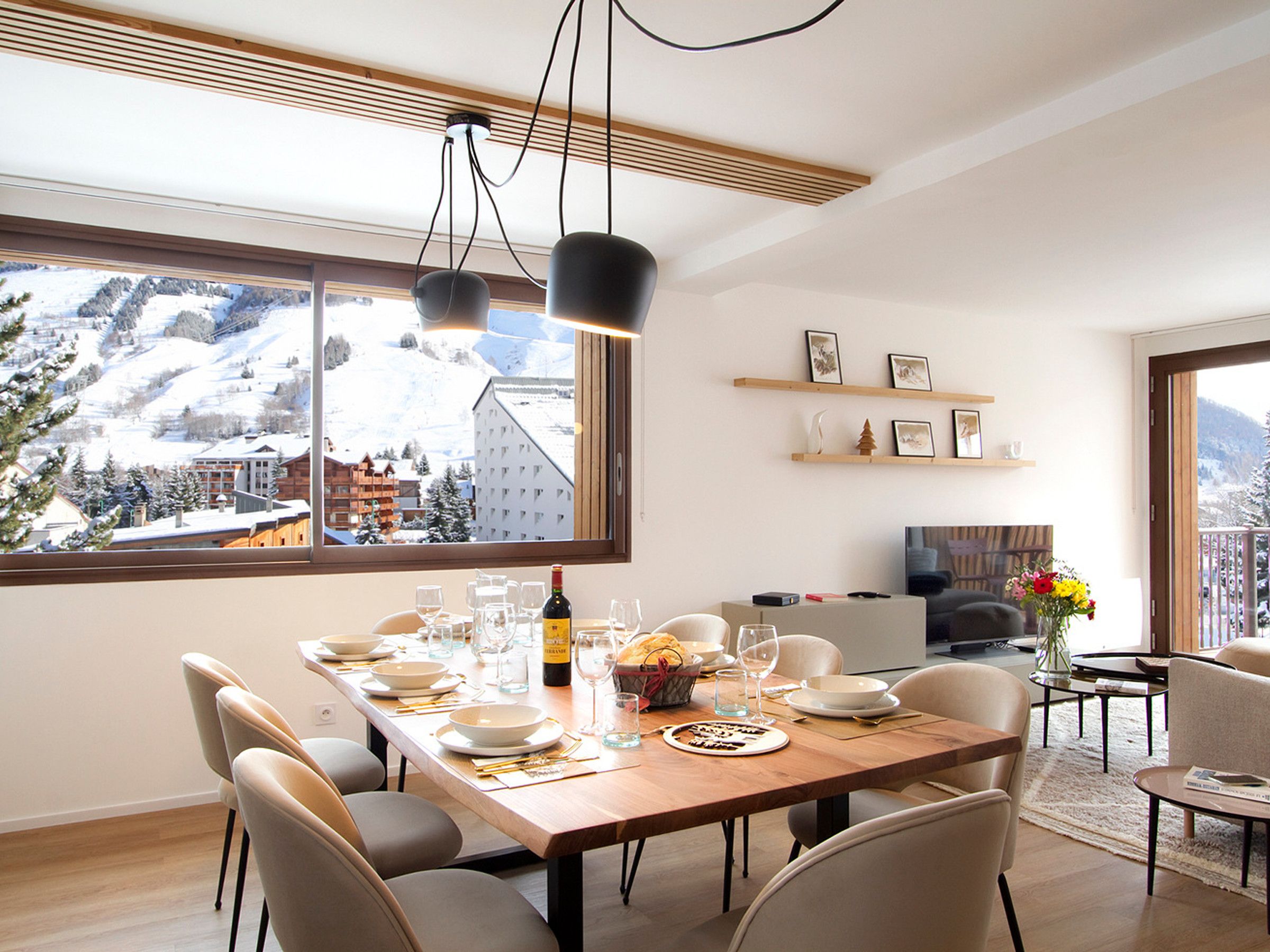 A dining area with a view of snowy mountains, a wooden table set for a meal, and a cozy living space with a TV and shelves.