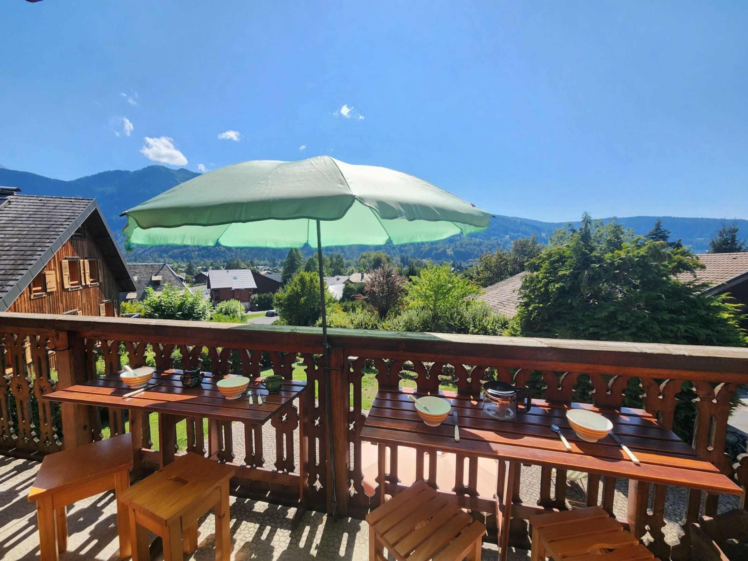 Une terrasse en bois avec des tables et des chaises, un parasol, et une vue sur des maisons et des montagnes en arrière-plan.