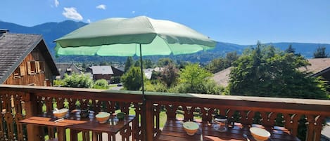 Une terrasse en bois avec des tables et des chaises, un parasol, et une vue sur des maisons et des montagnes en arrière-plan.