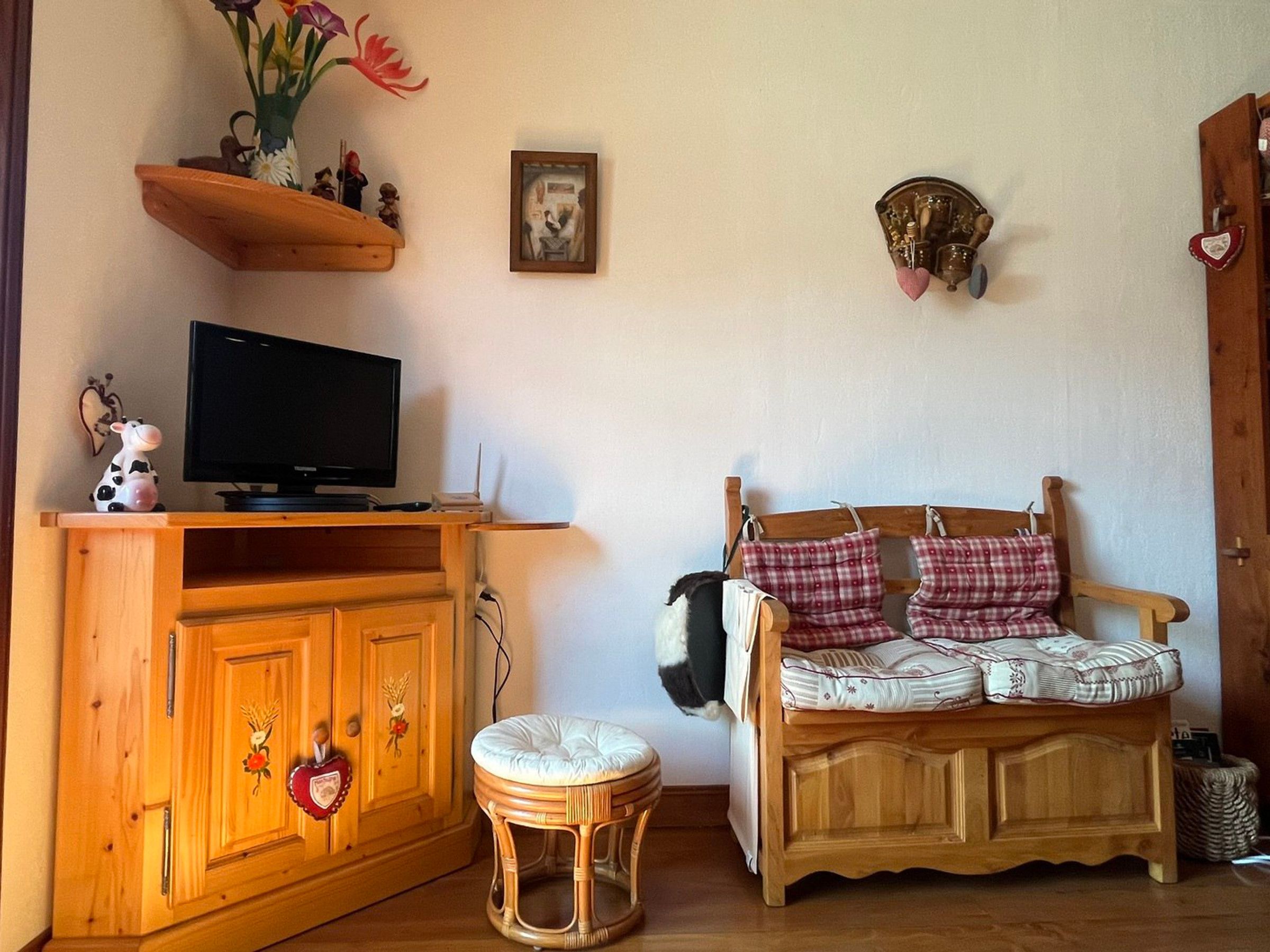 Een gezellige woonkamer met een houten kast, een televisie, een rieten krukje, een ruitjesbank en een decoratief object aan de muur.