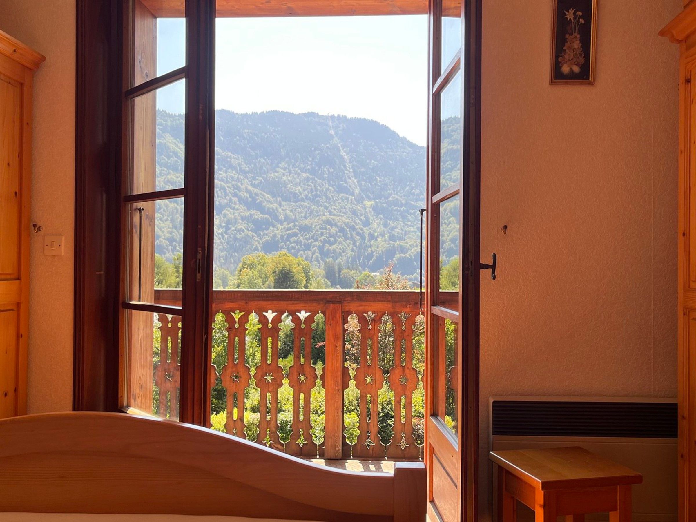 A room with a large window, a wooden bed, a small table, and a picture on the wall.