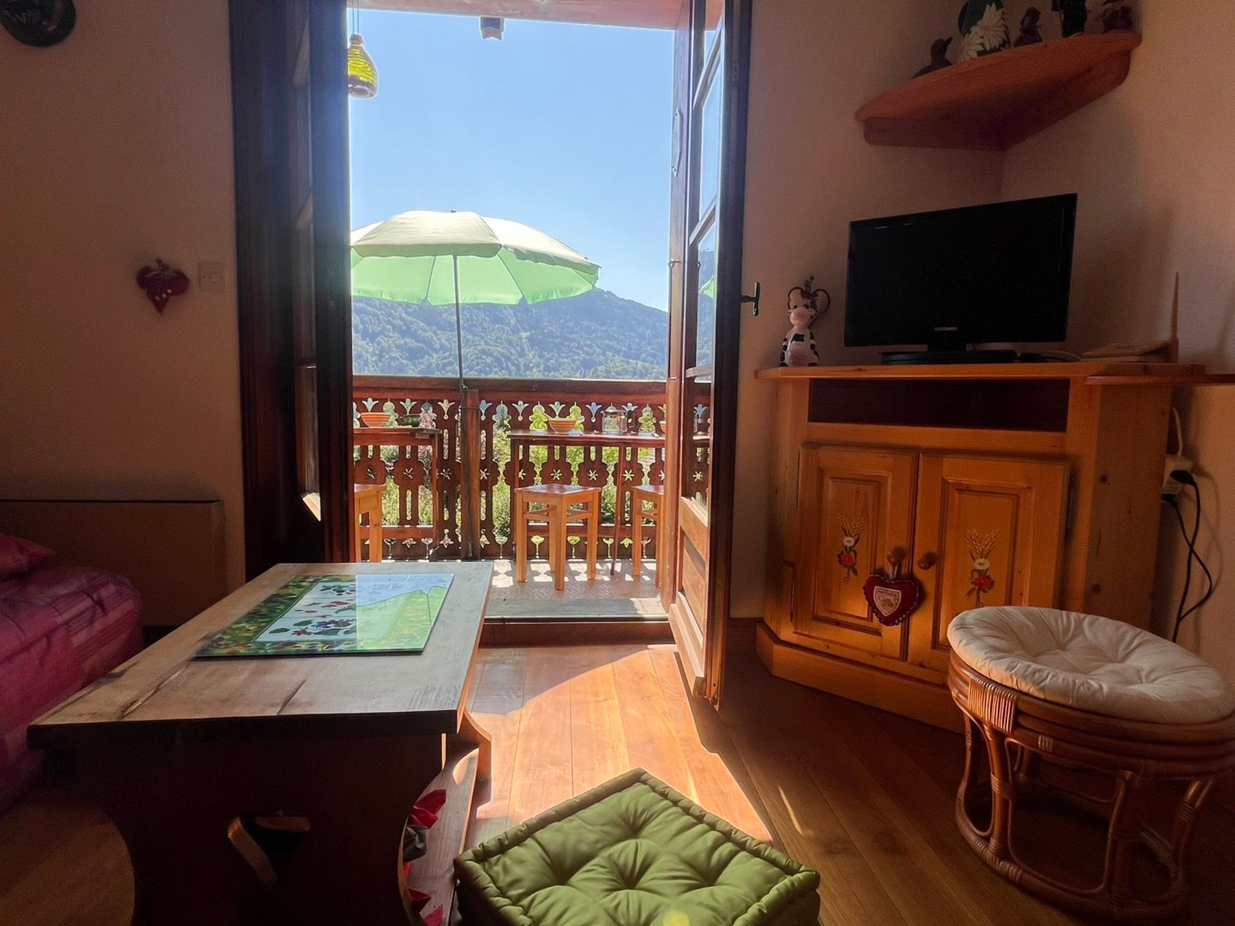 A room with a wooden table, a green cushioned seat, a television on a wooden cabinet, and a balcony with a view of mountains.