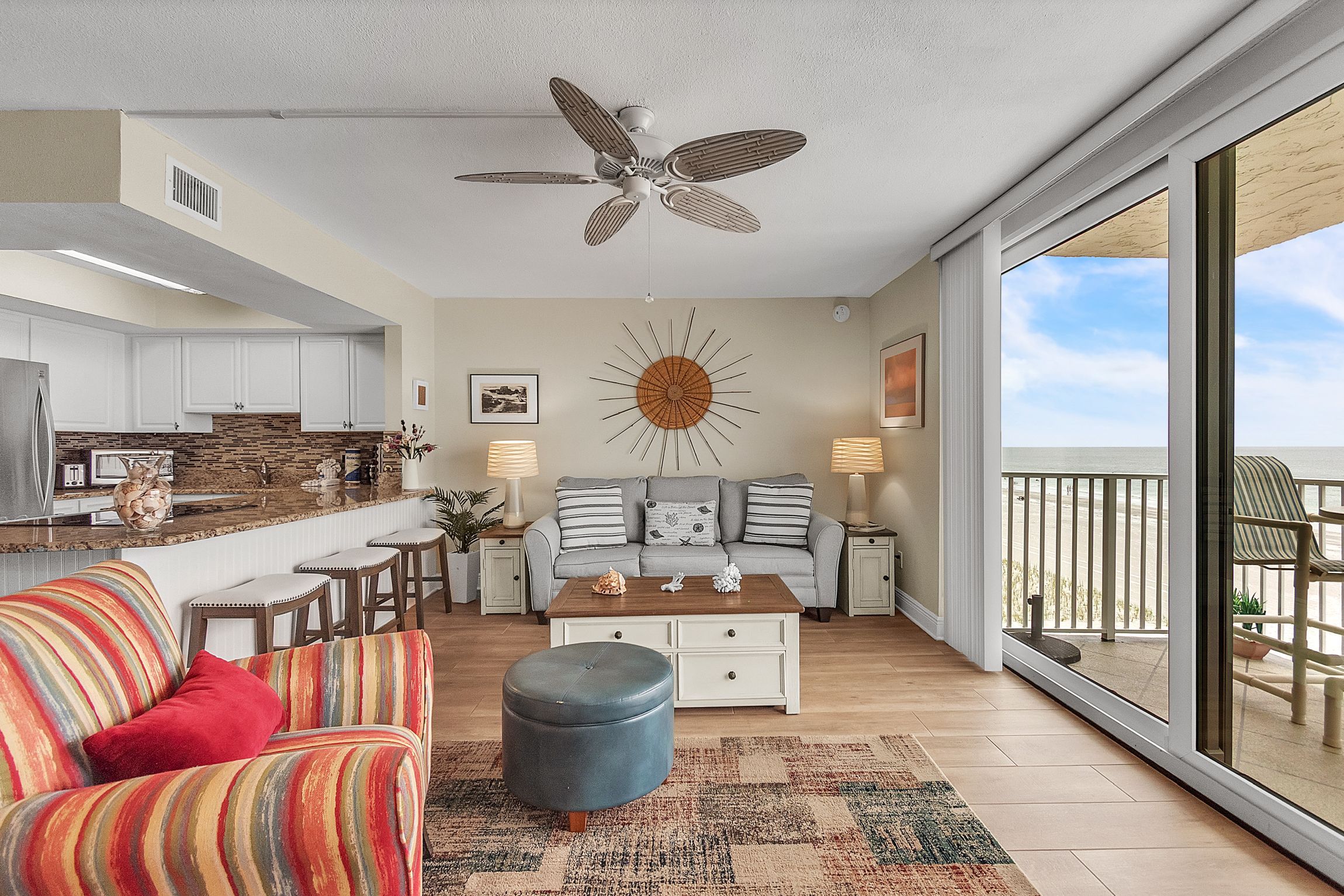 A spacious living area with a ceiling fan, a kitchen with white cabinets, a dining area with a gray sofa, and a balcony with a view.