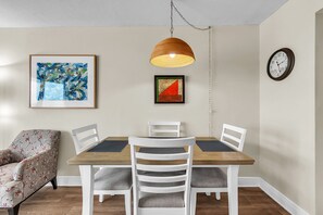 A dining area with a wooden table, white chairs, a hanging pendant light, and two framed artworks on the wall. - Beachfront Condo | Heated Pool | Private Balcony - Gulf Shores #404 (Indian Shores)