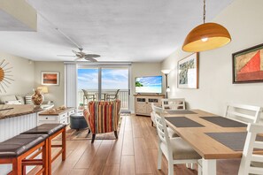 A spacious living area with a dining table, chairs, a sofa, a ceiling fan, and a view of the ocean through large windows. - Beachfront Condo | Heated Pool | Private Balcony - Gulf Shores #404 (Indian Shores)