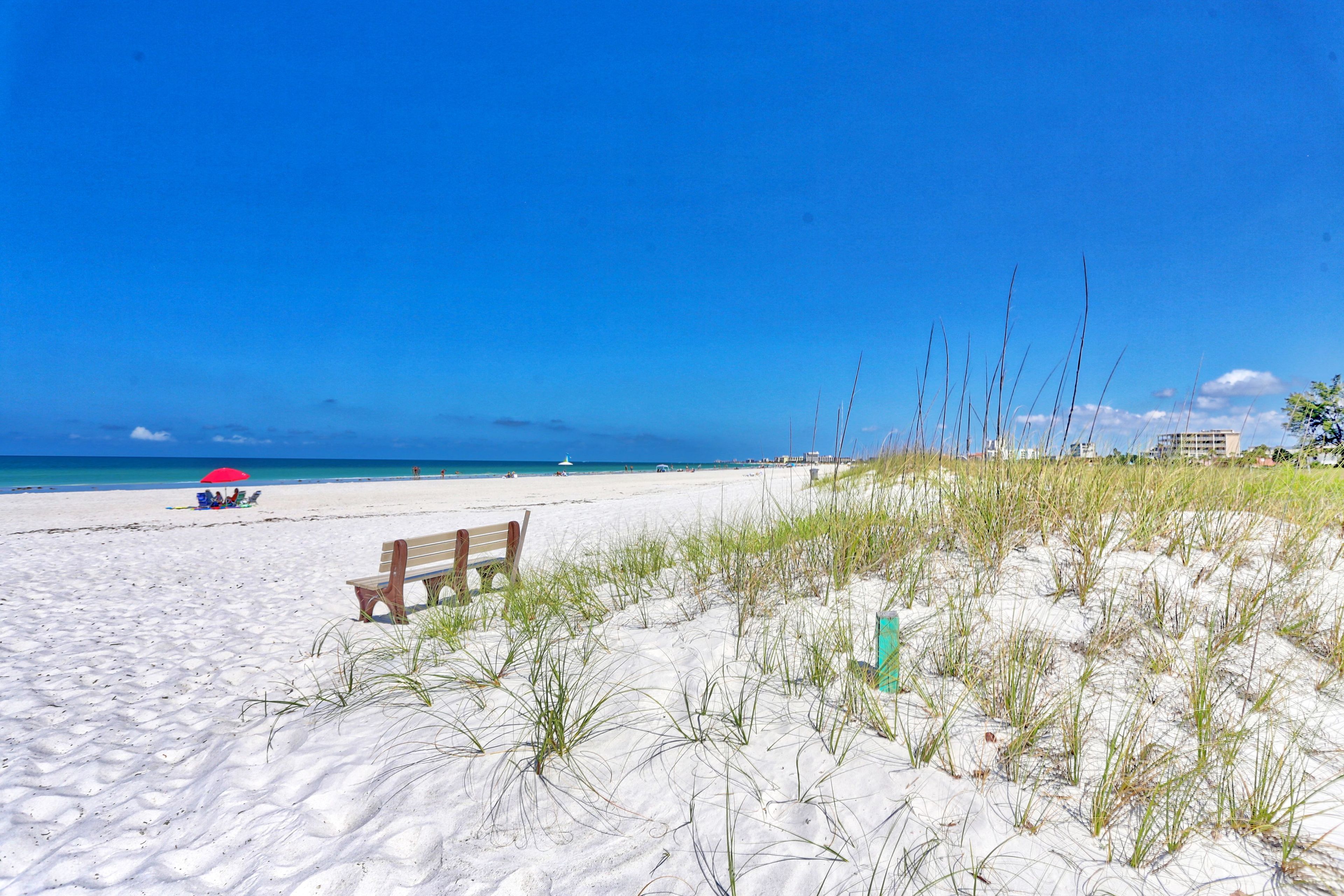 A sandy beach with a wooden bench and a red umbrella.