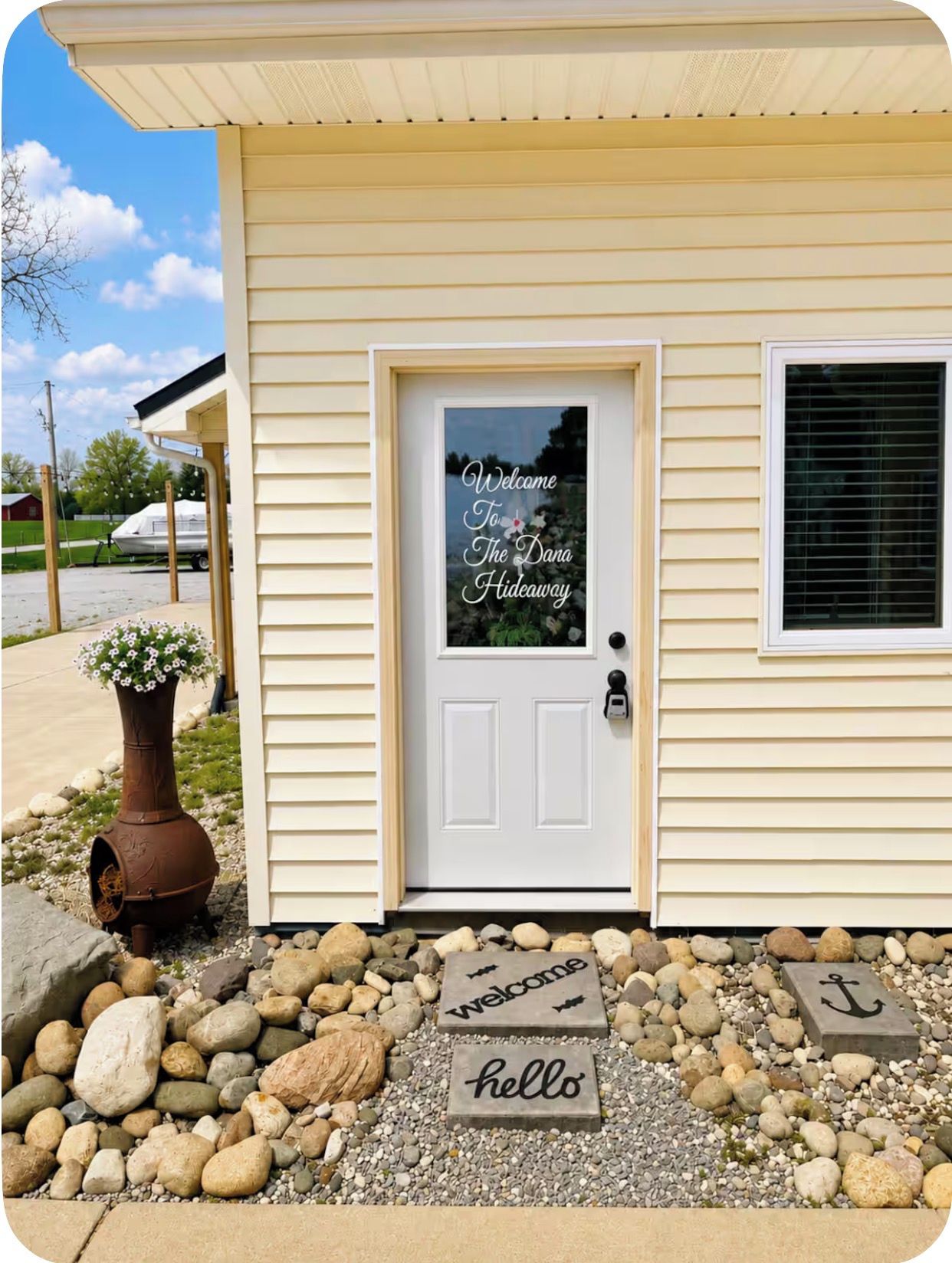 A house with a white door, a welcome sign, and a small garden with rocks and a flower pot.