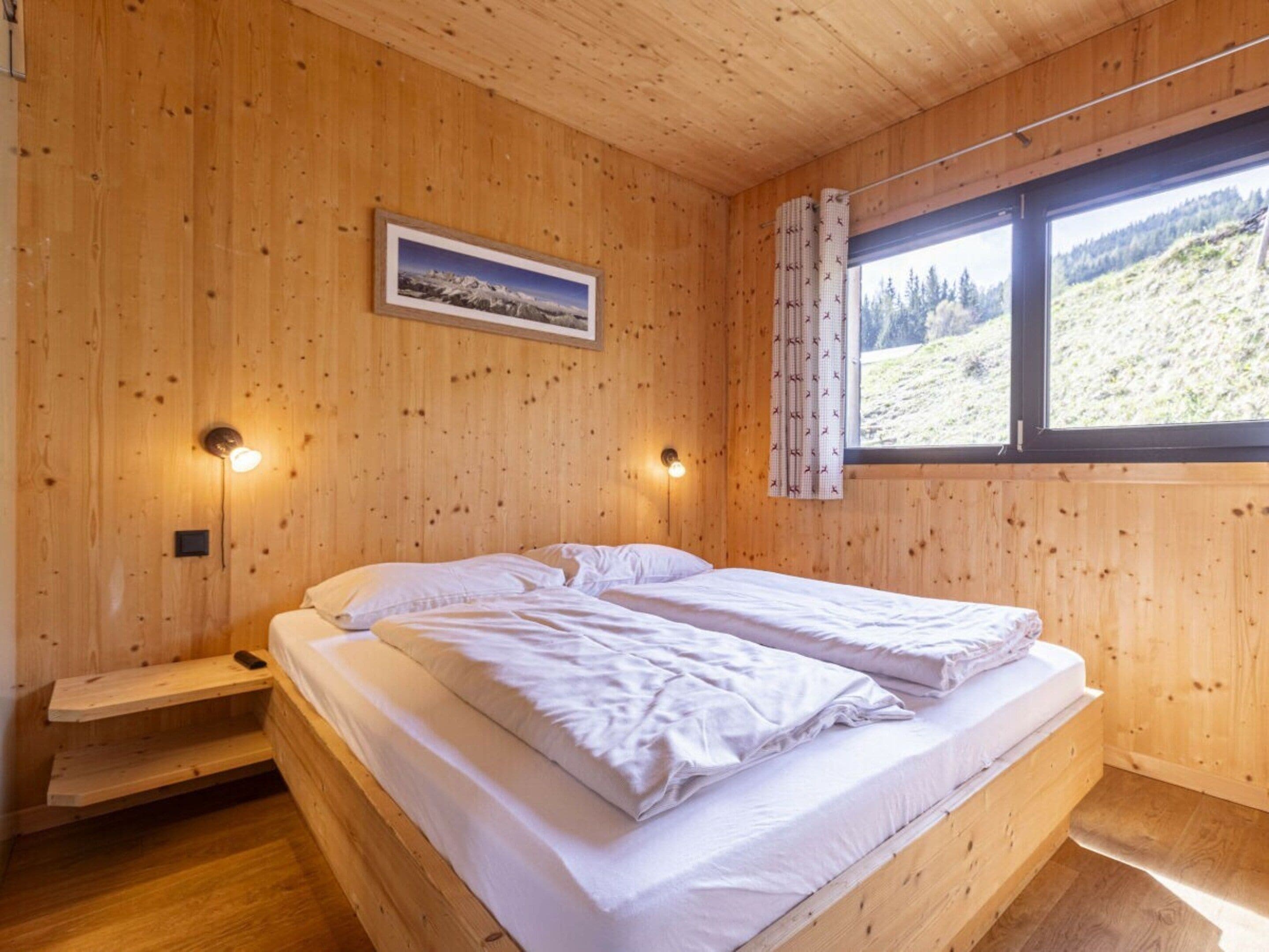 Une chambre de style cabane en bois, avec un lit, deux lampes fixées au mur, une fenêtre avec des rideaux et un tableau encadré accroché au mur.