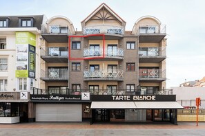 Een meerlaags gebouw met balkons, een winkel met een bordje 'Tarte Blanche' en een bord dat 'Zorgeloos' adverteert.