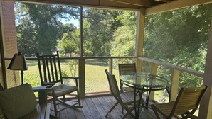 Uma área de estar coberta ao ar livre, com uma mesa, cadeiras, uma luminária e vista para um gramado.