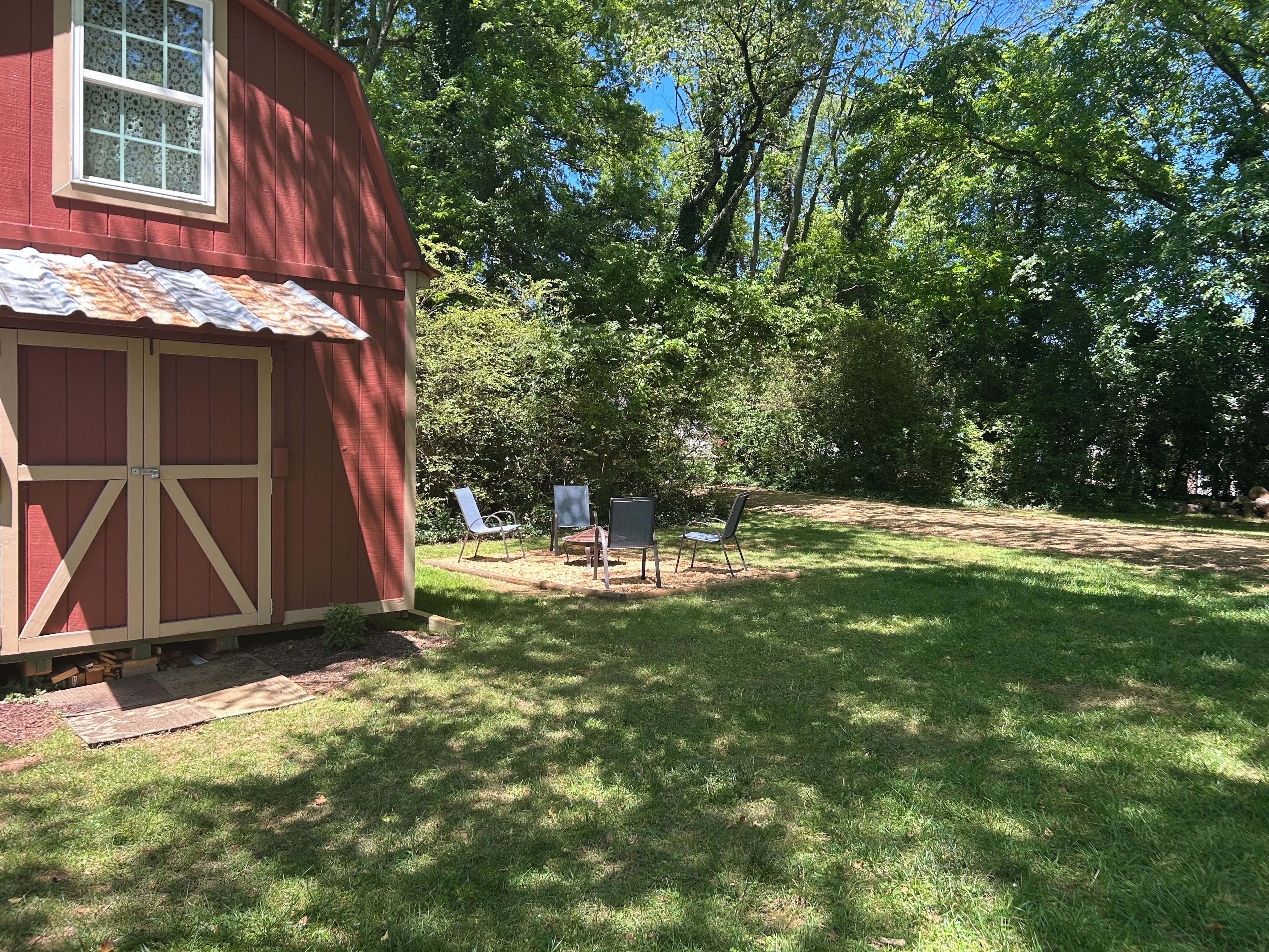 A red wooden shed with a window and a door, surrounded by a grassy yard with trees and a clear sky.