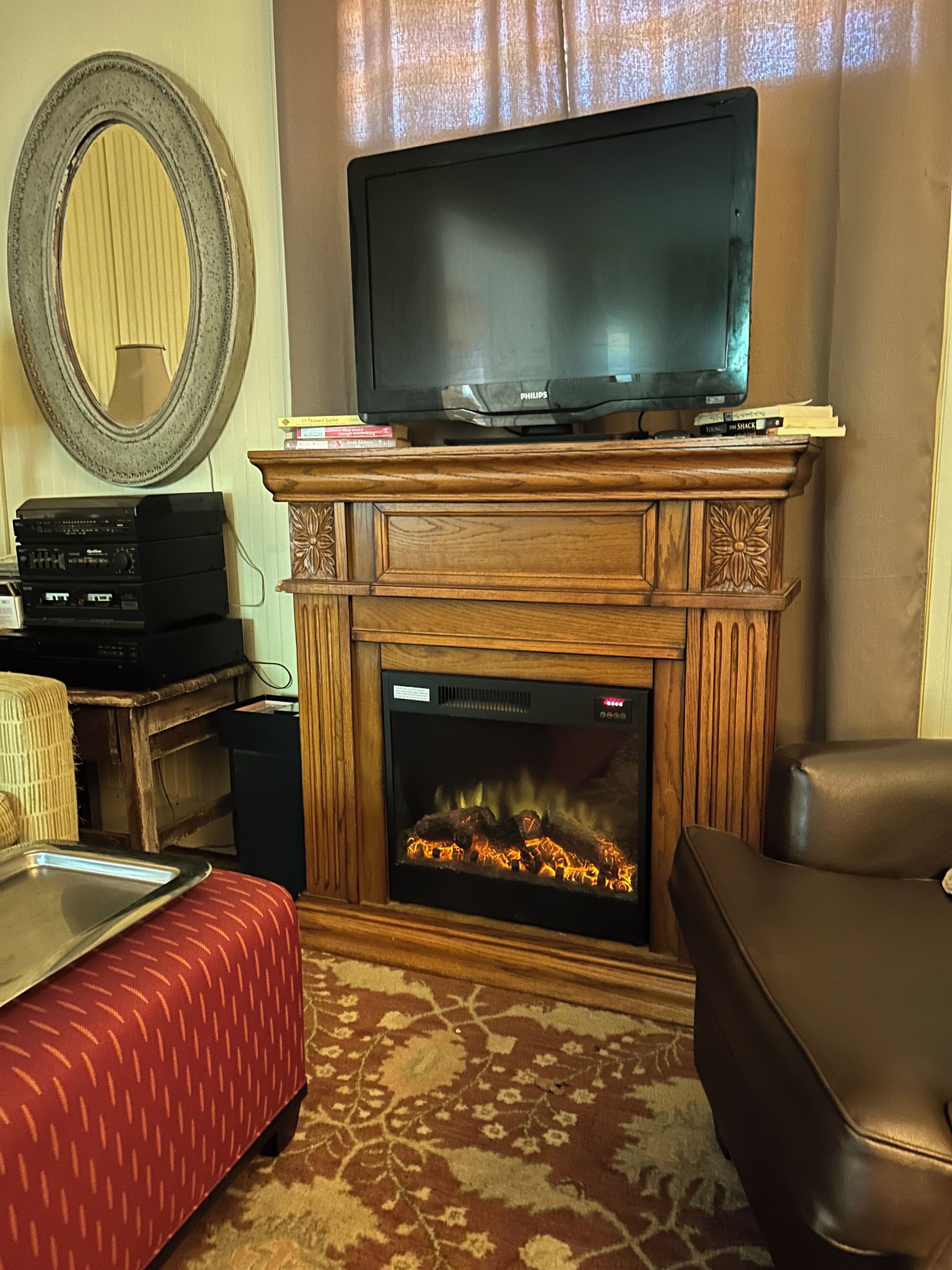 Ein Wohnzimmer mit einem Fernseher auf einem Holzständer, einem Kamin mit Feuer, einem roten, gemusterten Hocker und einem großen Spiegel an der Wand.
