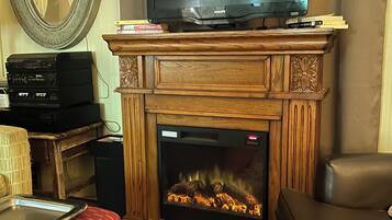 Ein Wohnzimmer mit einem Fernseher auf einem Holzständer, einem Kamin mit Feuer, einem roten, gemusterten Hocker und einem großen Spiegel an der Wand.