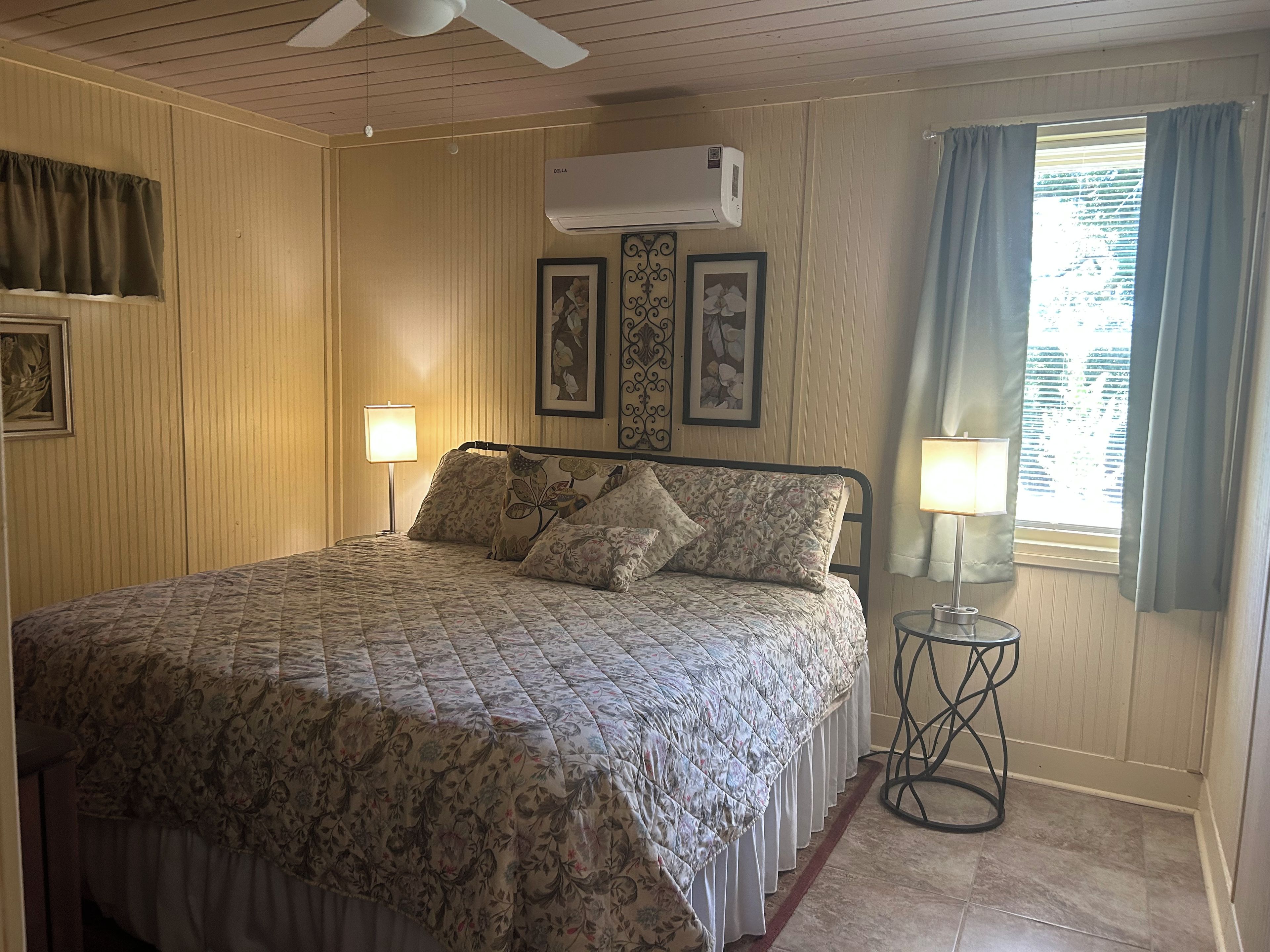 Quarto com cama, duas luminárias fixadas na parede, aparelho de ar condicionado e janela com cortinas.