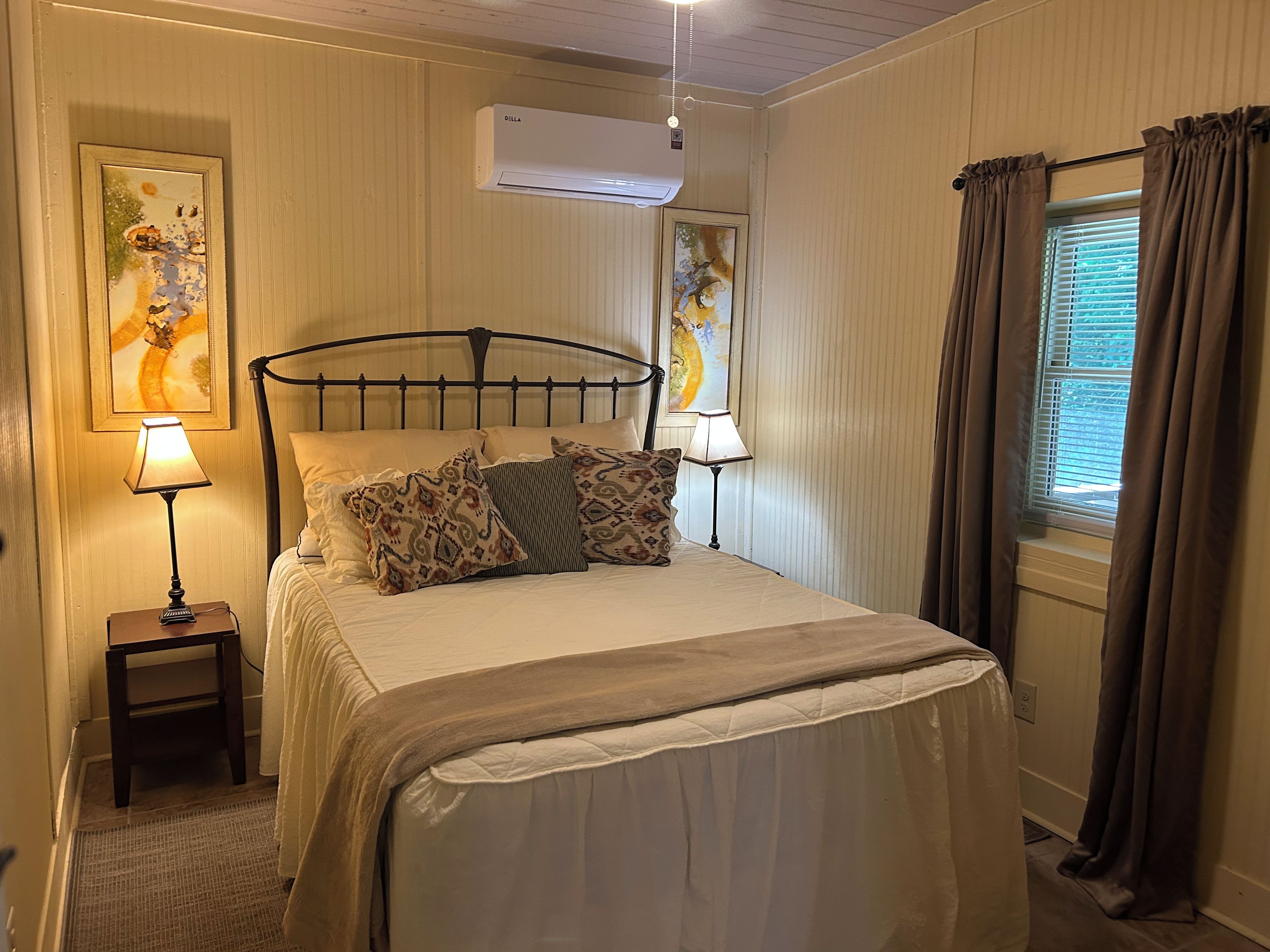 A neatly made bed with patterned pillows, a bedside lamp, a small nightstand, and a wall-mounted air conditioner.