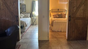 Quarto com cama, mesa de cabeceira e luminária. Uma porta dá acesso a outro cômodo.