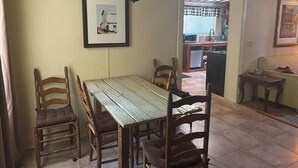 Uma sala de jantar com mesa e cadeiras de madeira, um quadro na parede e uma cozinha ao fundo.