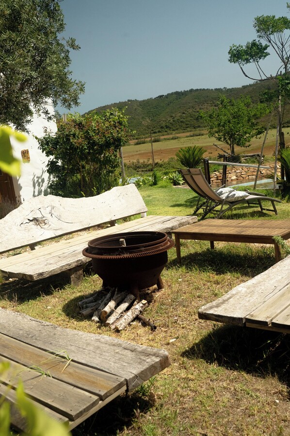 A rustic outdoor setting with a barbecue pit, wooden lounge chairs, and a white building in the background.