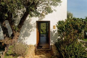 A doorway leading to a covered outdoor area with a bench and a chair, surrounded by greenery and a stone wall.
