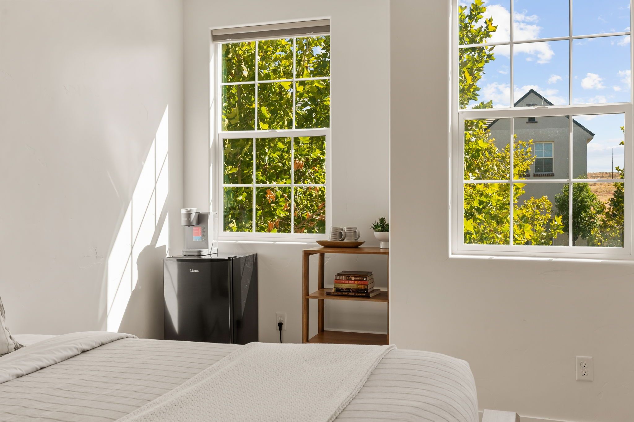 A bedroom with a bed, a coffee machine, a shelf with books, and two windows with a view of trees and a house.